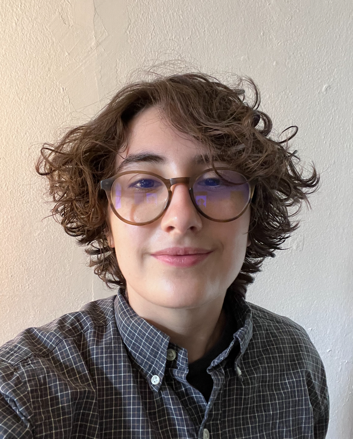 Student portrait of a person with short, curly brown hair, wearing glasses and a plaid shirt, smiling subtly.