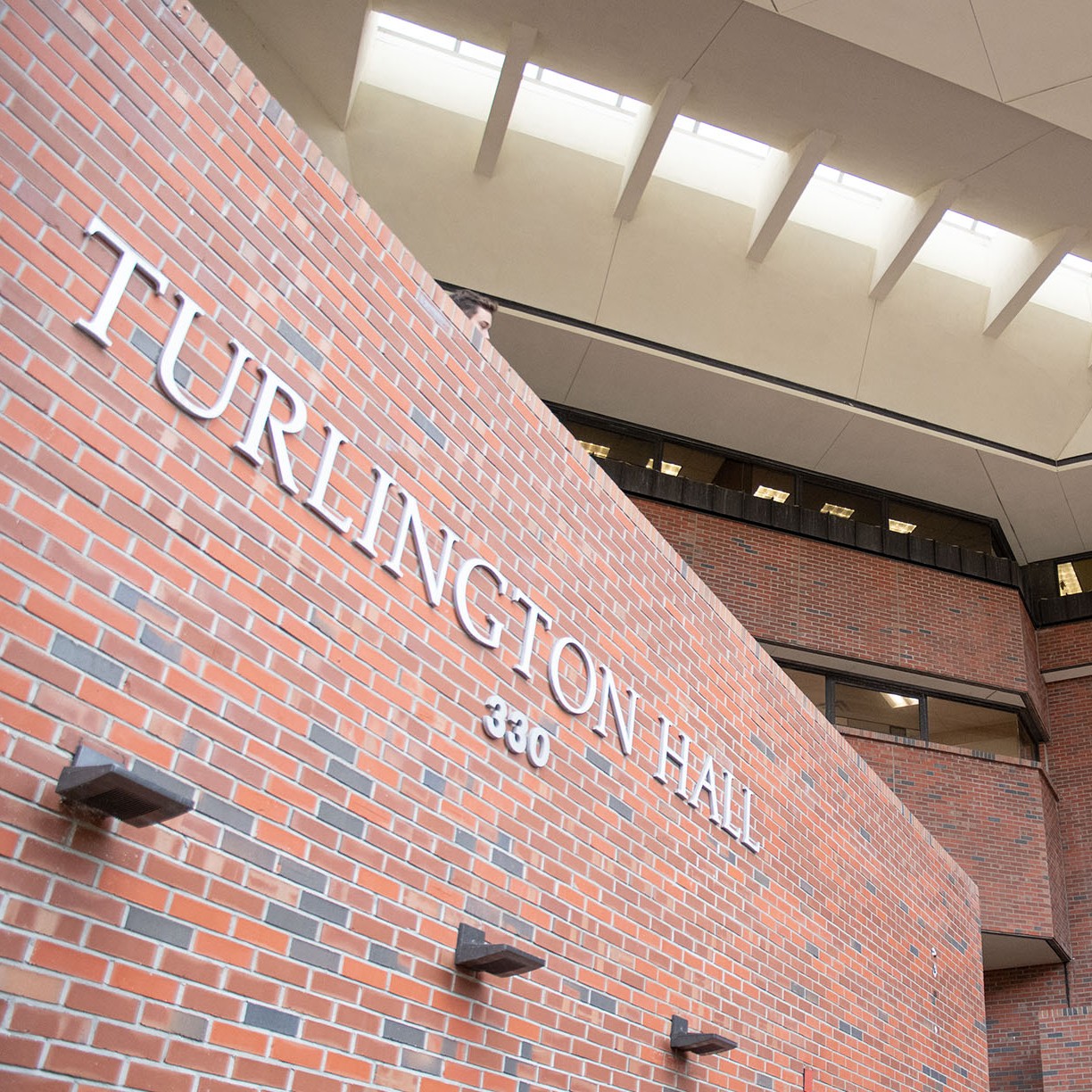 Photograph of a brick building exterior featuring a large wall with "TURLINGTON HALL 330" in white letters. The ceiling has skylights allowing natural light, and multiple floors with windows and balconies are visible in the background. Photographed by Tricia Lopez